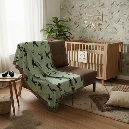 A mint green woven throw blanket with a black bird pattern and fringed edges, draped over a brown armchair against a white background.